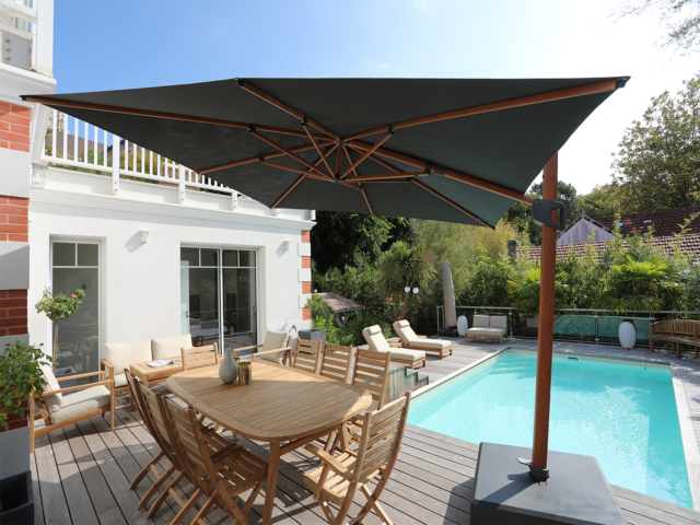 Parasol déporté – Centaurus Sooty aspect teck 3x3m avec sa toile gris Sooty et sa structure aspect teck. Le parasol est accompagné d'une table fixe ovale en Teck et de chaises et fauteuils en Teck Nos structures évoluent : en inclinant les mâts à 6° vers l’arrière nous avons gagné en stabilité en reculant le centre de gravité du milieu du parasol vers le pied du parasol. Nous avons également ajouté des amortisseurs en bout de baleines.